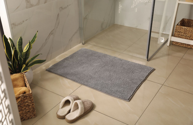 Un tapis de bain gris texturé posé sur un carrelage beige clair devant une douche en verre,