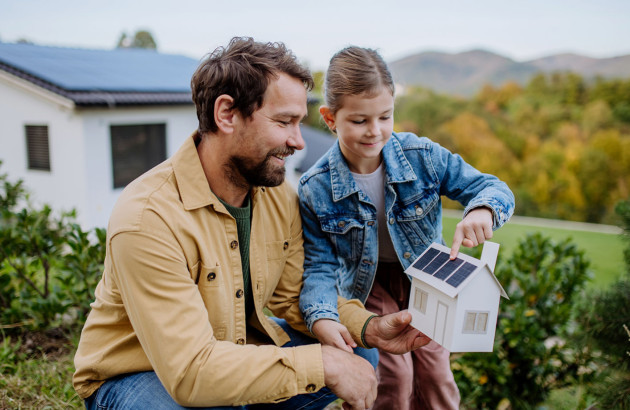 Père de famille qui explique à sa fille ce qu'est un panneau solaire sur une maquette de maison